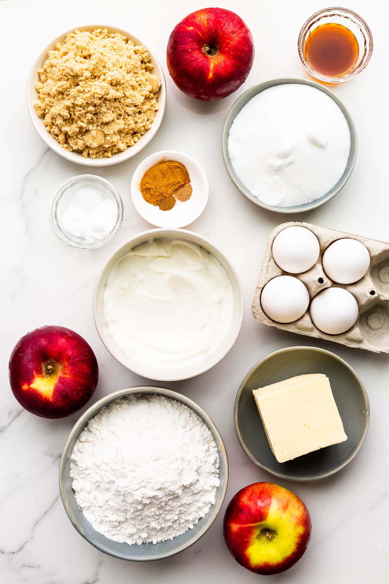 Ingredients to make apple bundt cake measured out including apples, brown sugar, white sugar, spices, sour cream, eggs, salt, baking powder, baking soda, butter, and flour