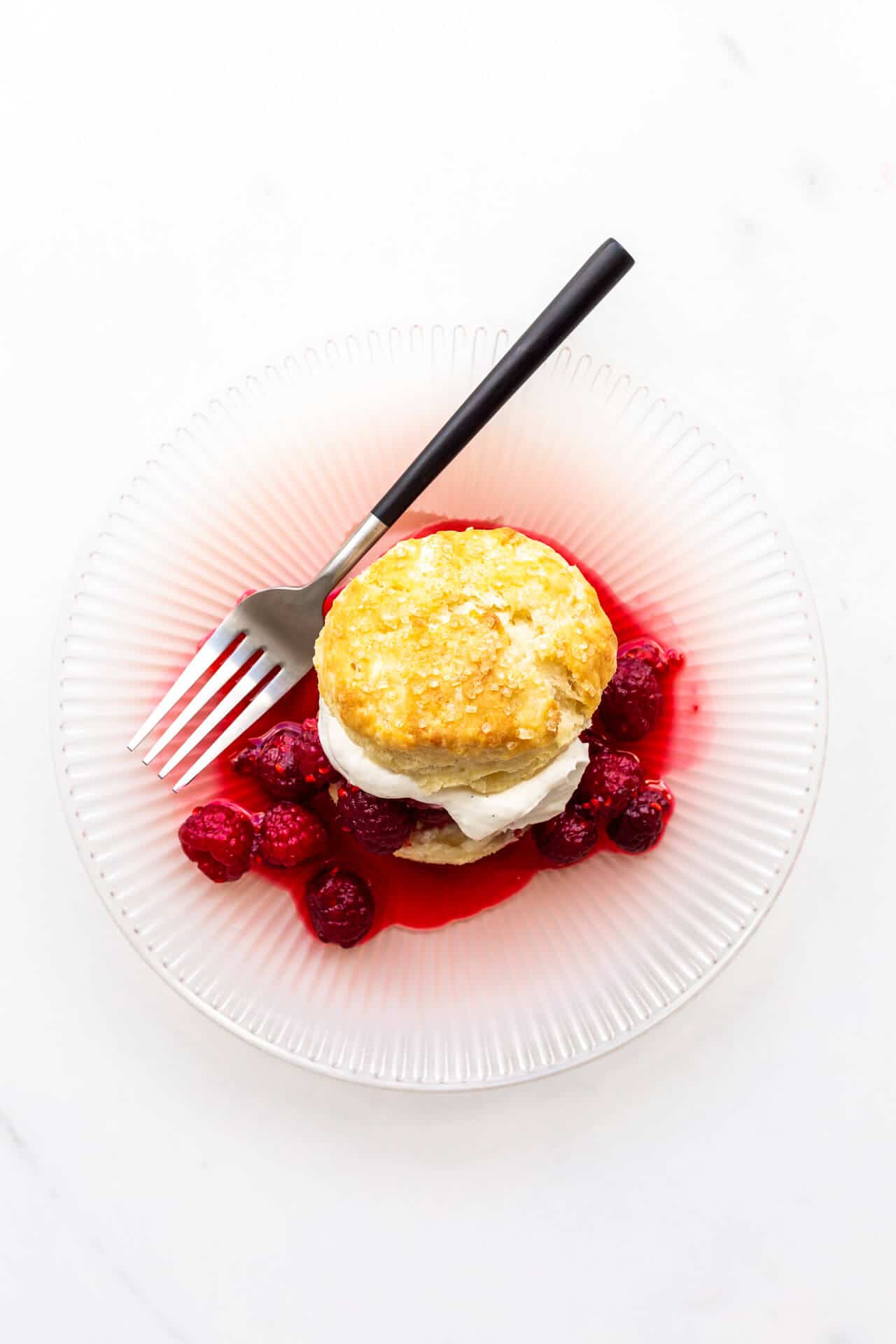 A raspberry shortcake with homemade biscuit, whipped cream, and berries on a pink plate with a fork with black matte handle