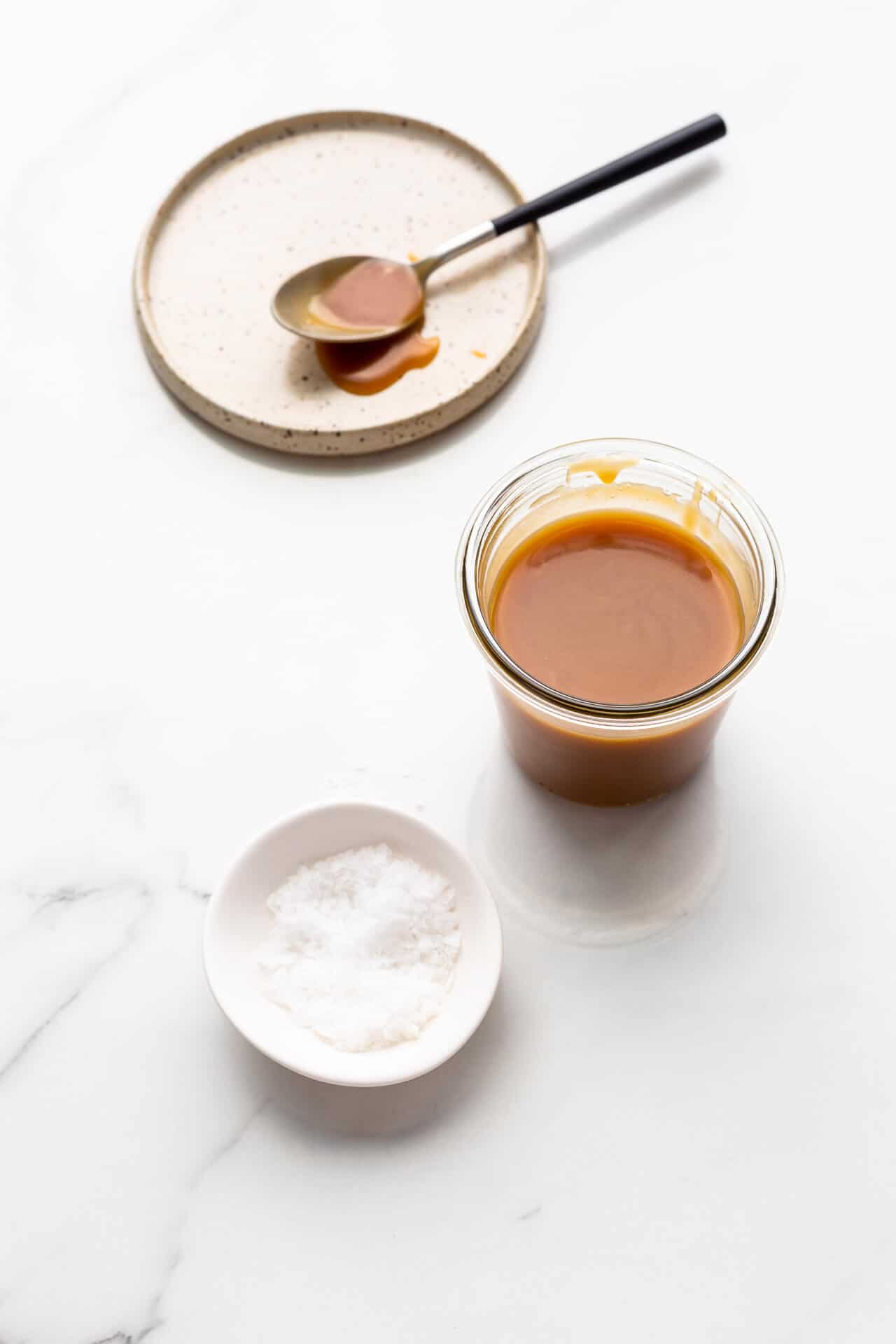 Salted caramel in a jar with a small bowl of flaky sea salt and a ceramic plate in the background with a spoon of caramel on it