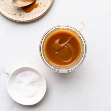 Homemade salted caramel sauce in a jar with a little pinching bowl of sea salt and a ceramic plate with a spoon with salted caramel on it