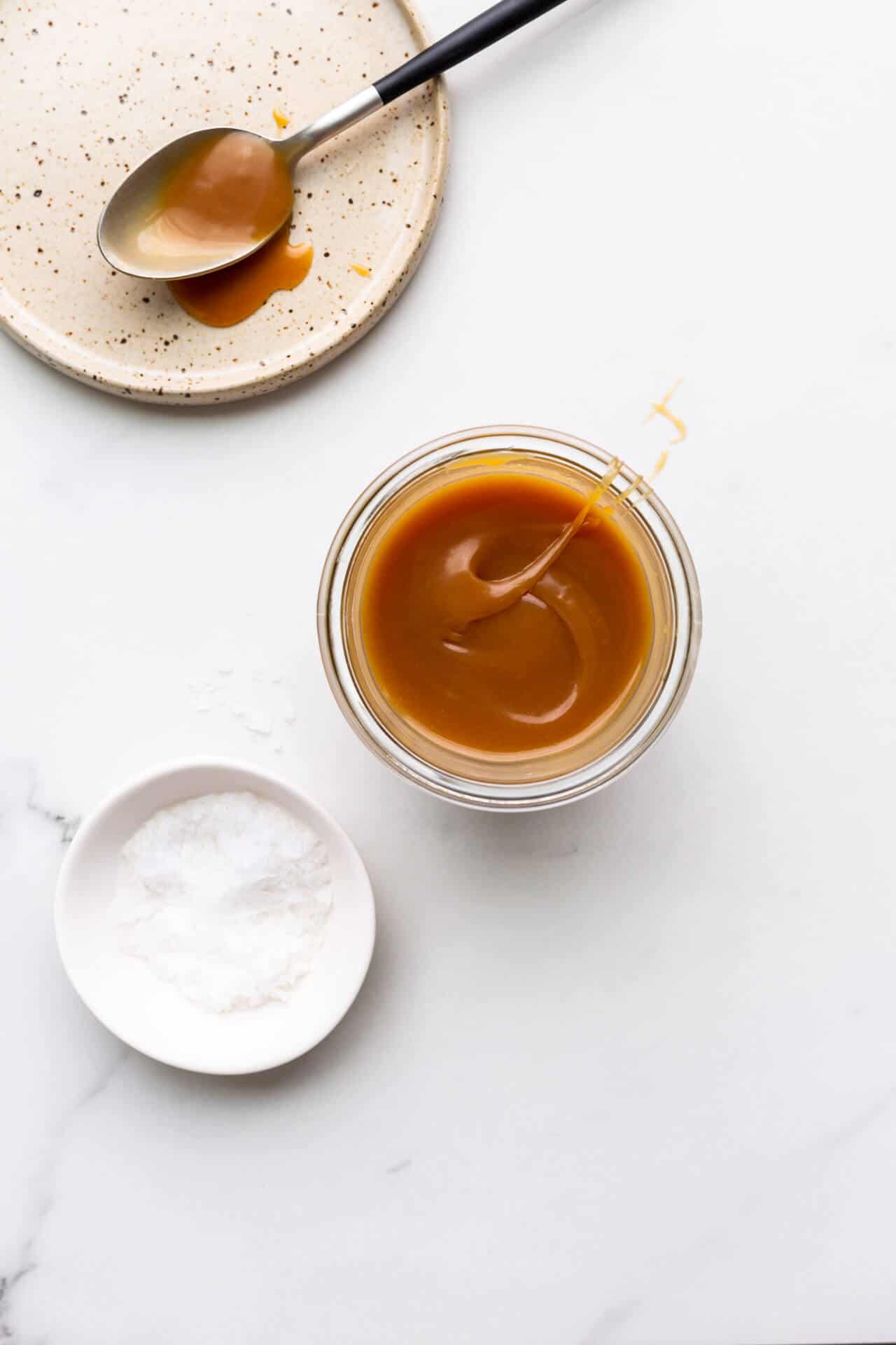 Homemade salted caramel sauce in a jar with a little pinching bowl of sea salt and a ceramic plate with a spoon with salted caramel on it