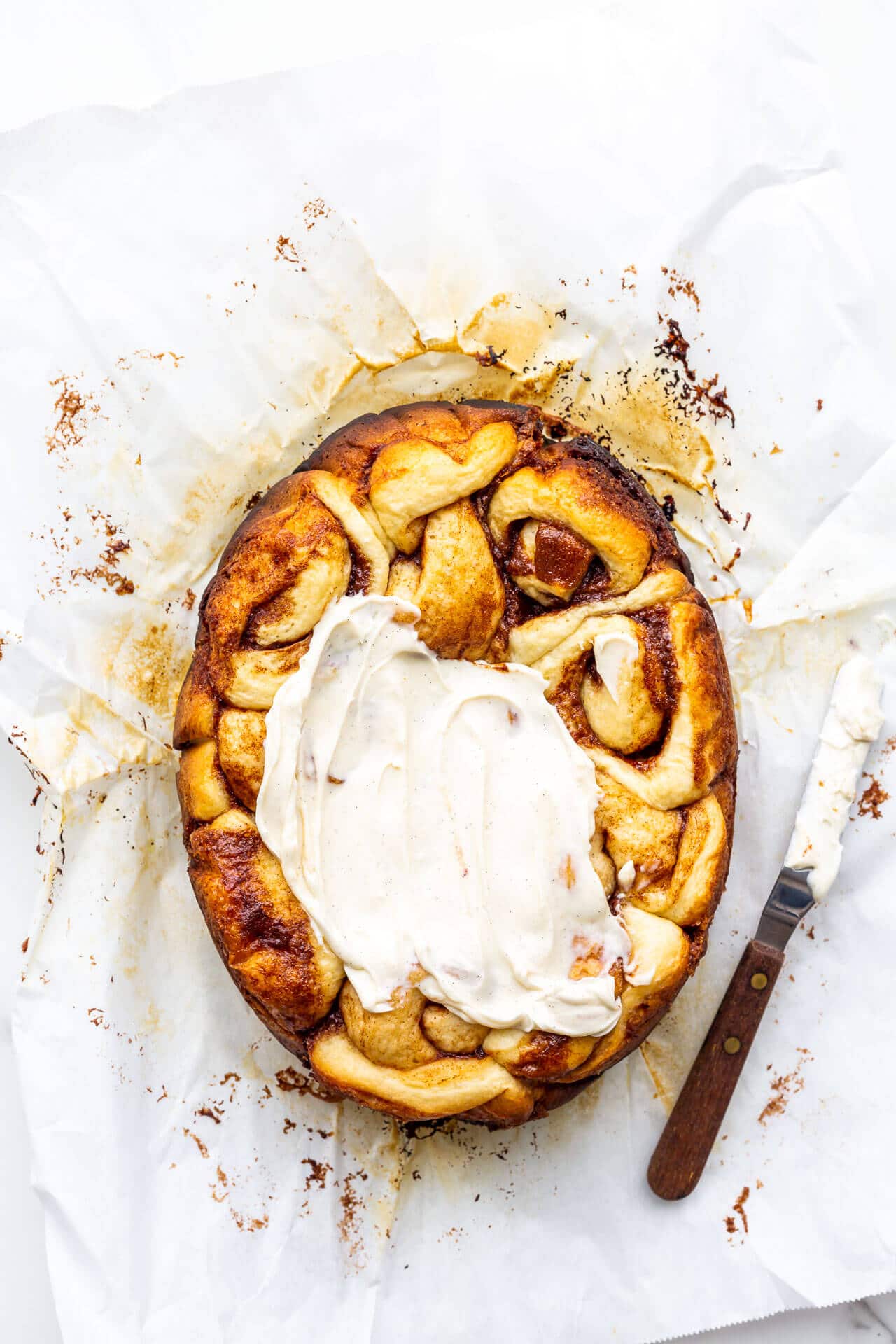 Cinnamon buns on parchment paper being frosted with cream cheese frosting using an offset spatula