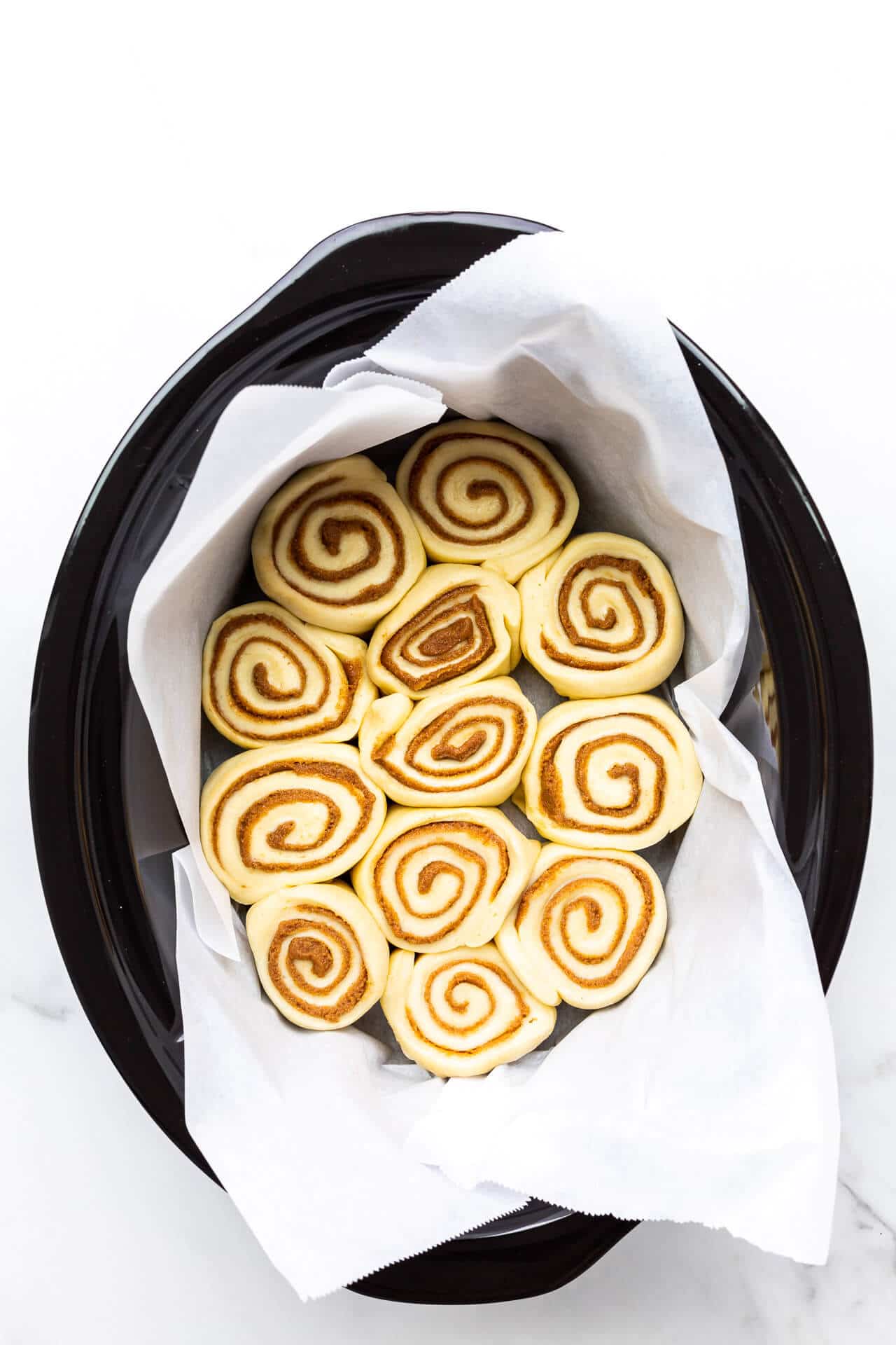 Cinnamon buns placed in a parchment-lined slow cooker black ceramic insert ready to be baked