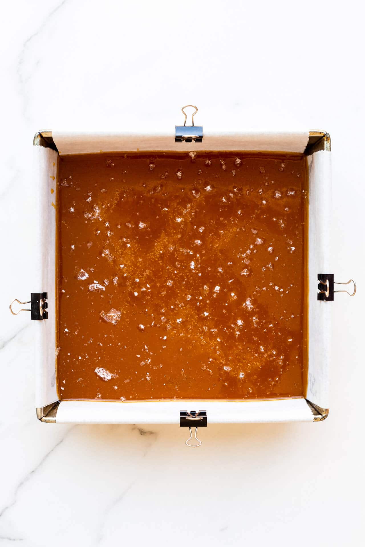 Caramels setting in an 8x8 pan lined with parchment paper clipped to sides of pan to hold it in place