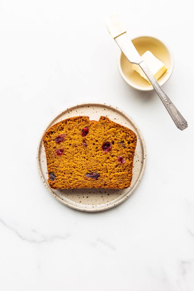 A slice of pumpkin cranberry bread on a speckled plate with a little bowl of butter and a knife