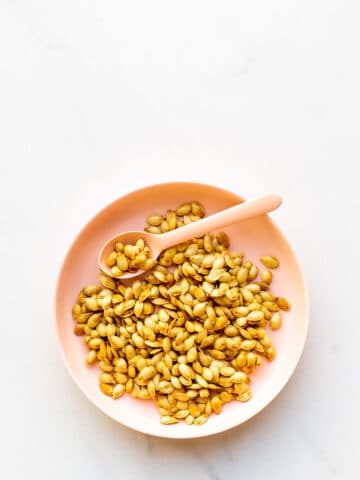 A pink bowl of toasted pumpkin seeds with a pink spoon for serving