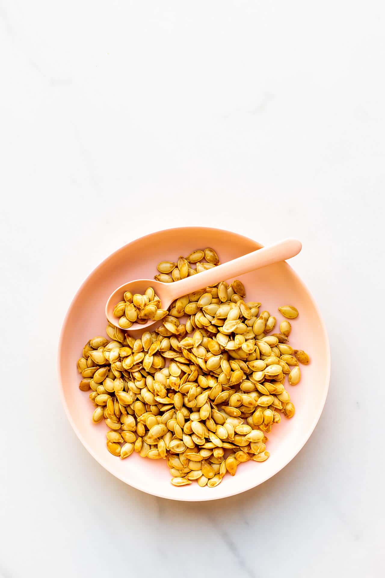 A pink bowl of toasted pumpkin seeds with a pink spoon for serving