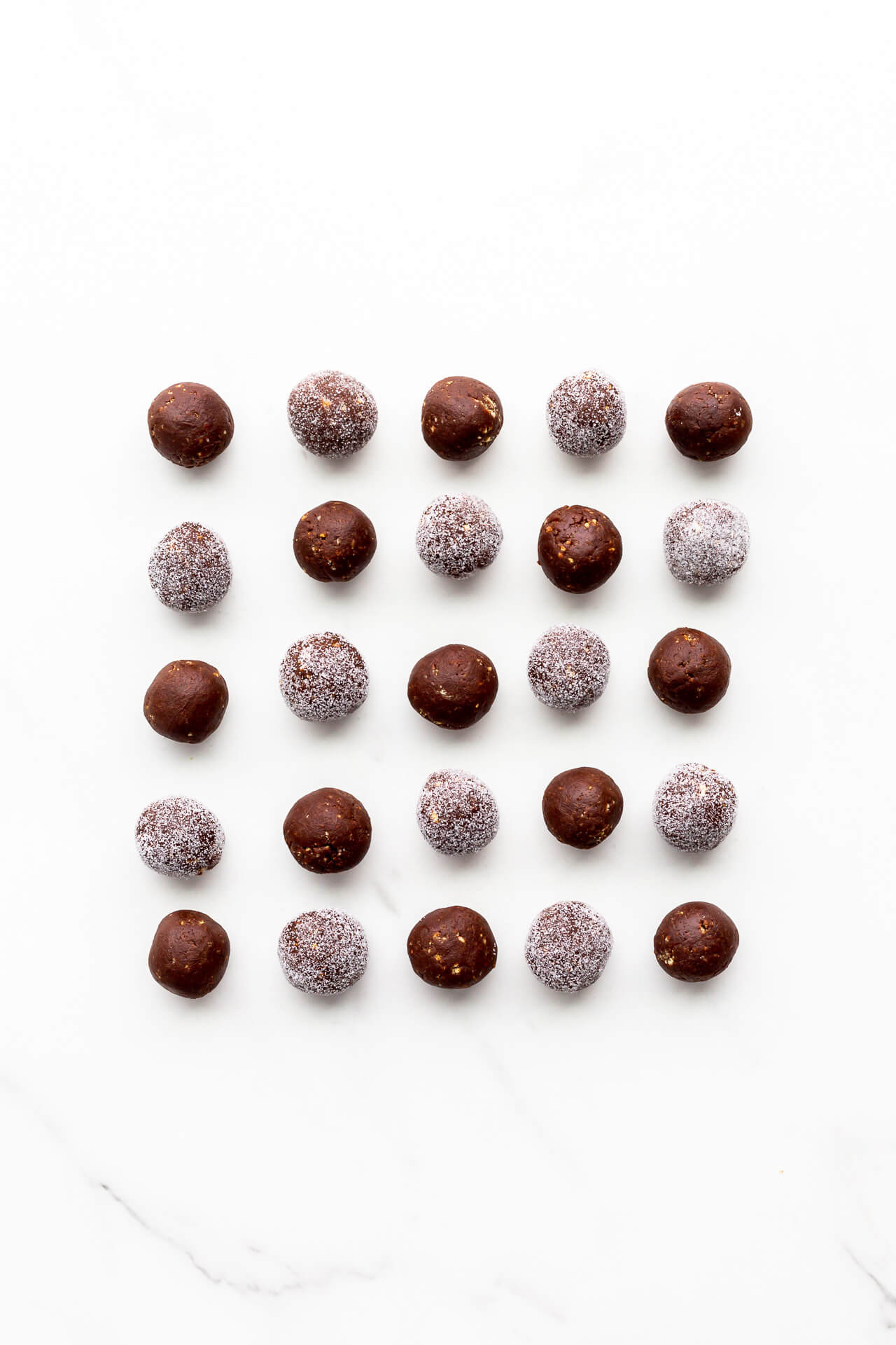 Homemade chocolate amaretto booze balls arranged on a marble surface in a grid pattern, some coated with granulated sugar and some not.