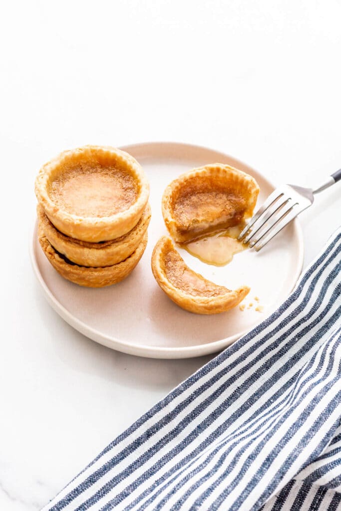 A beige plate with a stack of three butter tarts and a fourth next to it, cut open with a fork to reveal the gooey filling.