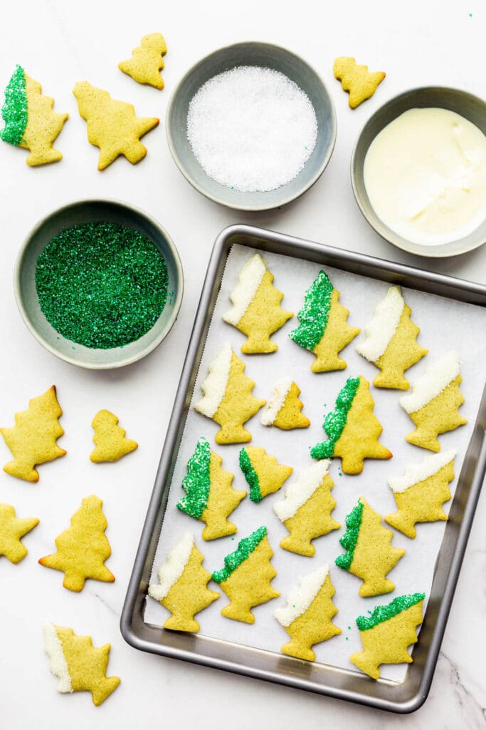 Dipping matcha green tea gingerbread cookie cutouts shaped like trees in white chocolate and coloured sugar sprinkles.