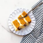 A slice of carrot cake layer cake with cream cheese frosting on a pale blue plate with a fork and striped linen napkin.
