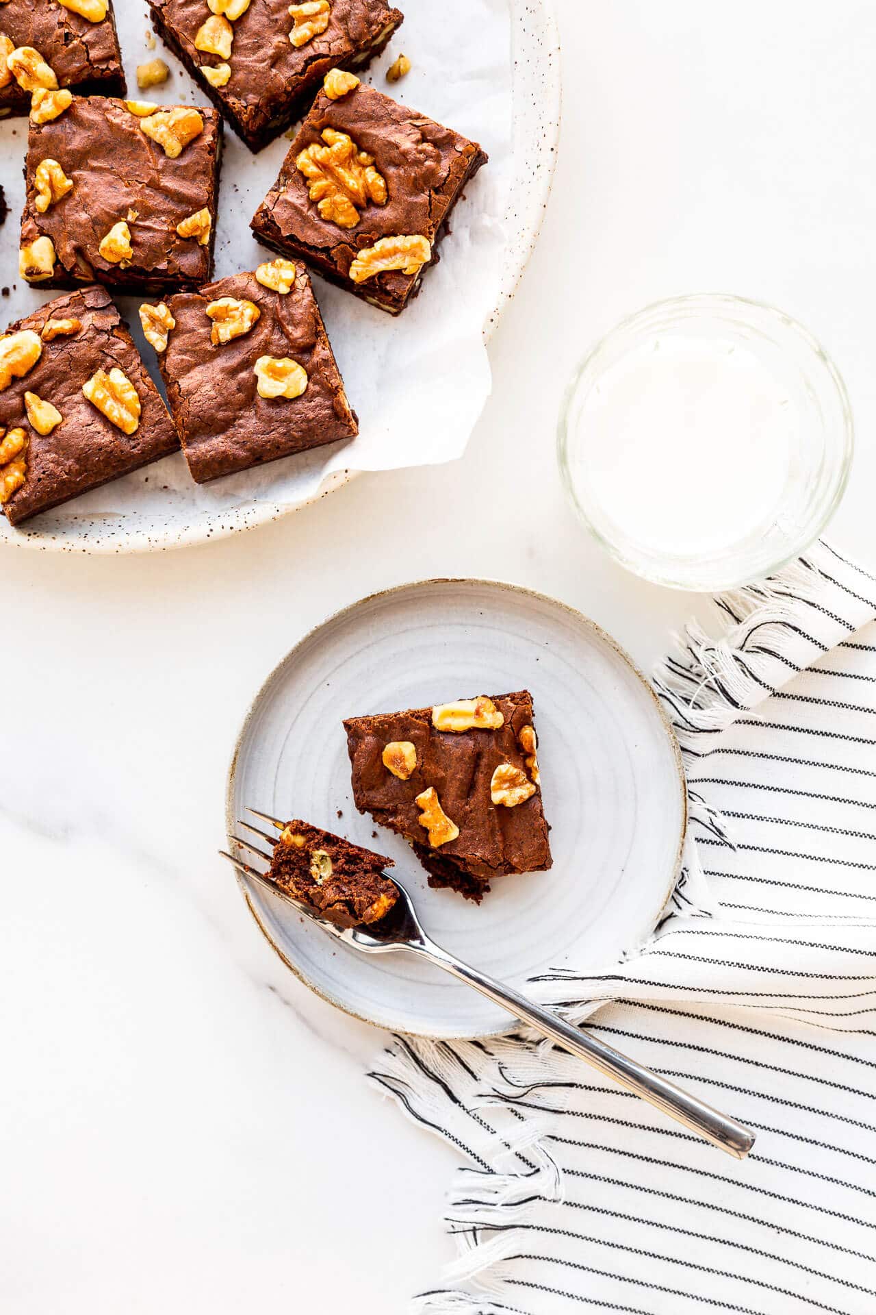 A plate of brownies with walnuts with one set on a dessert plate with a glass of milk.