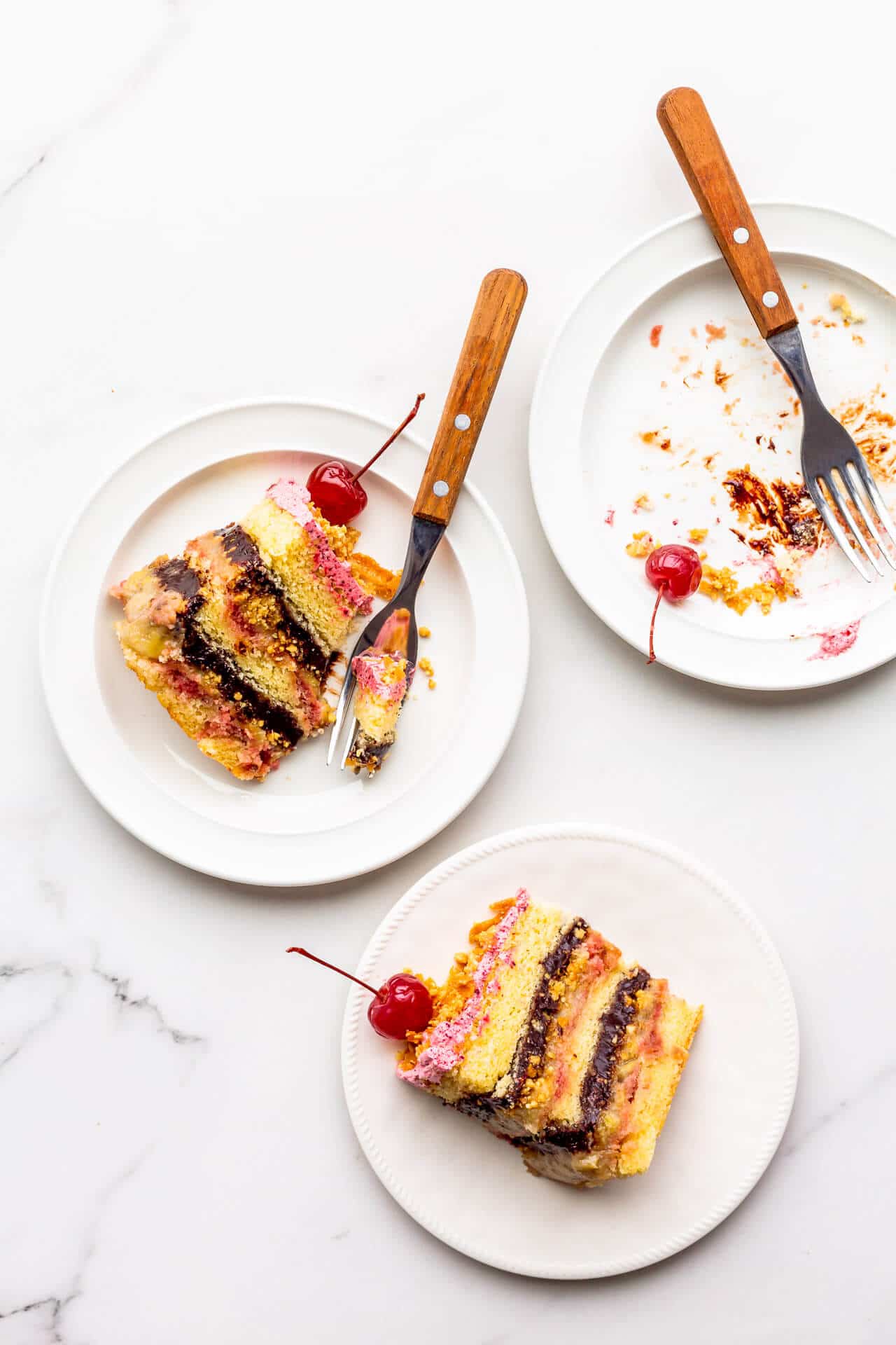 Slices of banana split cake topped with maraschino cherries, served on plates, with one slice eaten.