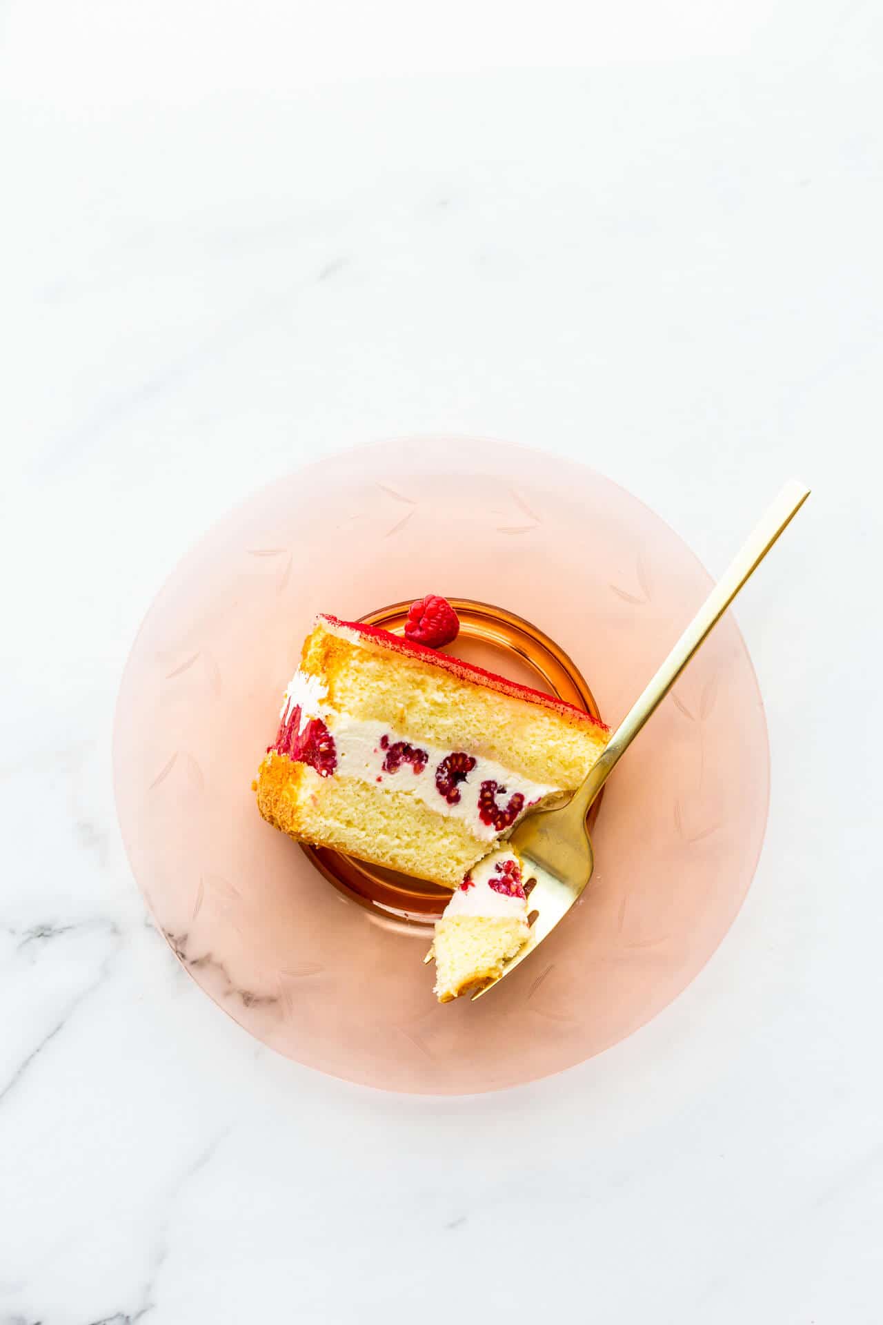 A slice of framboisier cake on a pink glass plate with a gold fork.