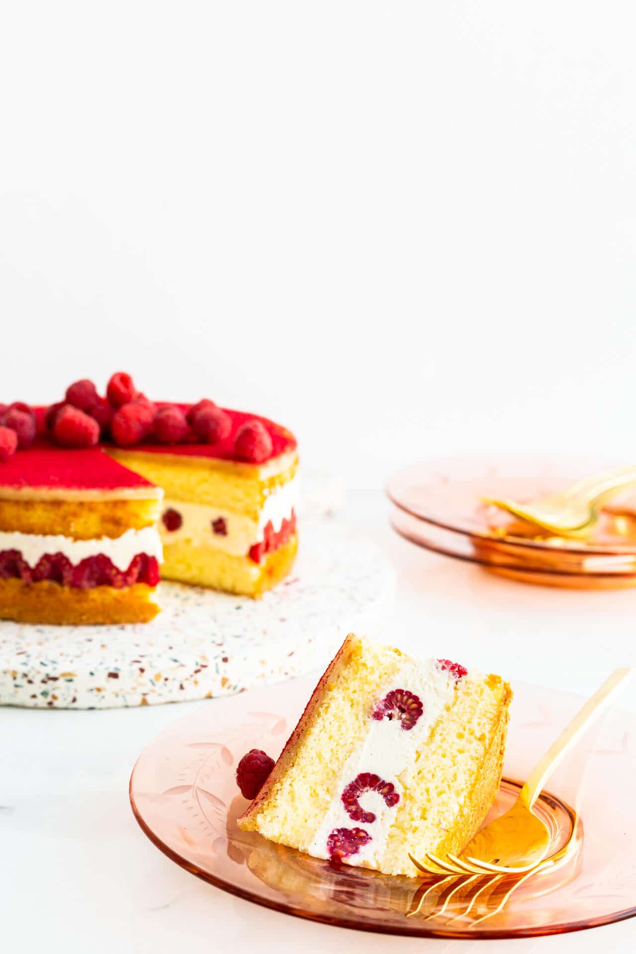 Framboisier cake topped with fresh raspberries on a terrazzo board being served on pink glass plates.