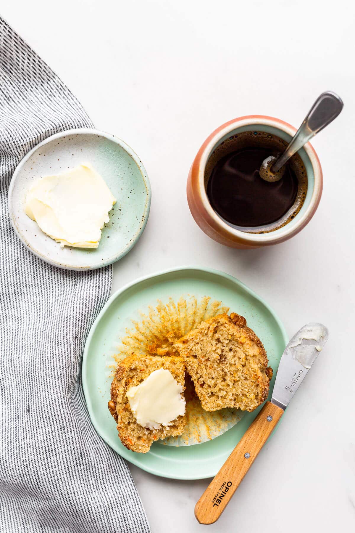 Freshly baked carrot muffin on a plate, split open and buttered. Served with a cup of coffee.