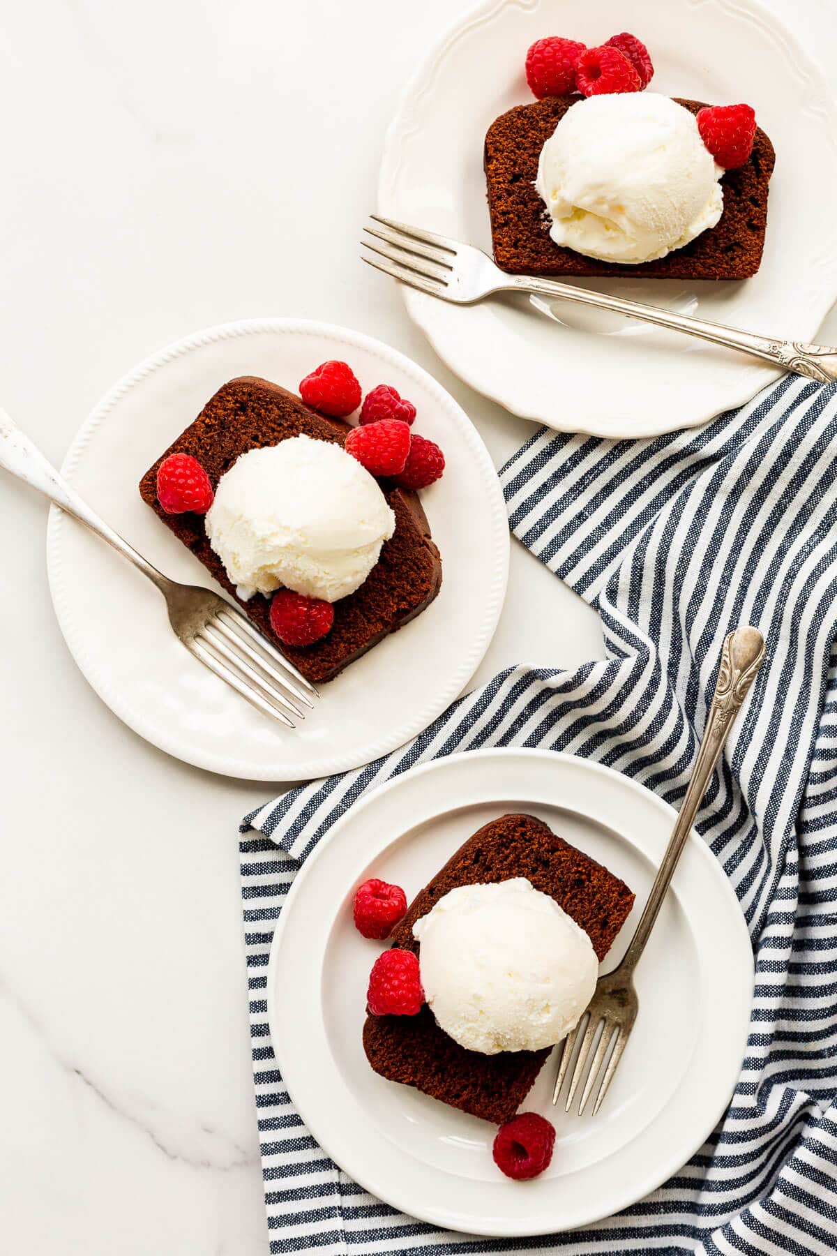 Slice of chocolate loaf cake with a scoop of ice cream and fresh raspberries on a plate with a fork, with a striped linen tucked underneath.