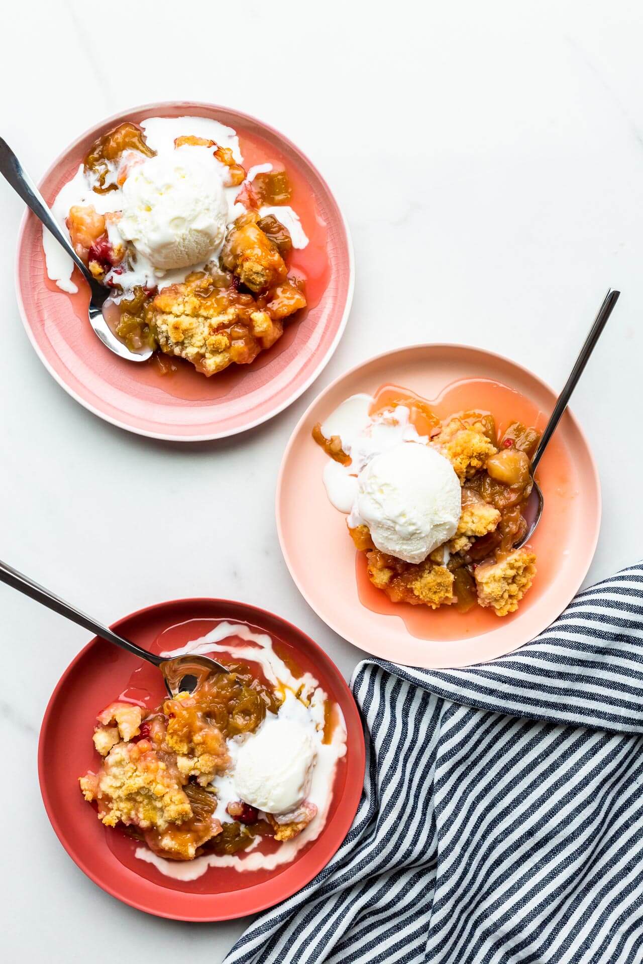 Three pink bowls of homemade rhubarb crisp served warm with scoops of vanilla ice cream melting on top.