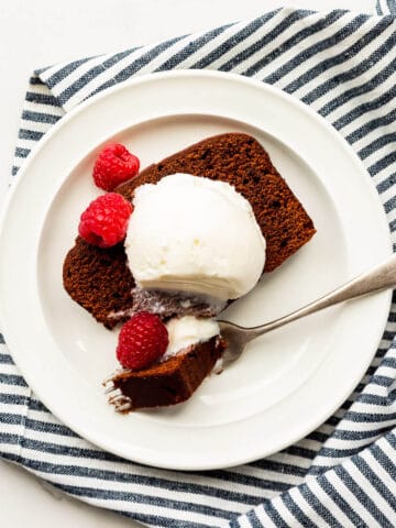 Slice of chocolate loaf cake with a scoop of ice cream and fresh raspberries on a plate with a fork, with a striped linen tucked underneath.