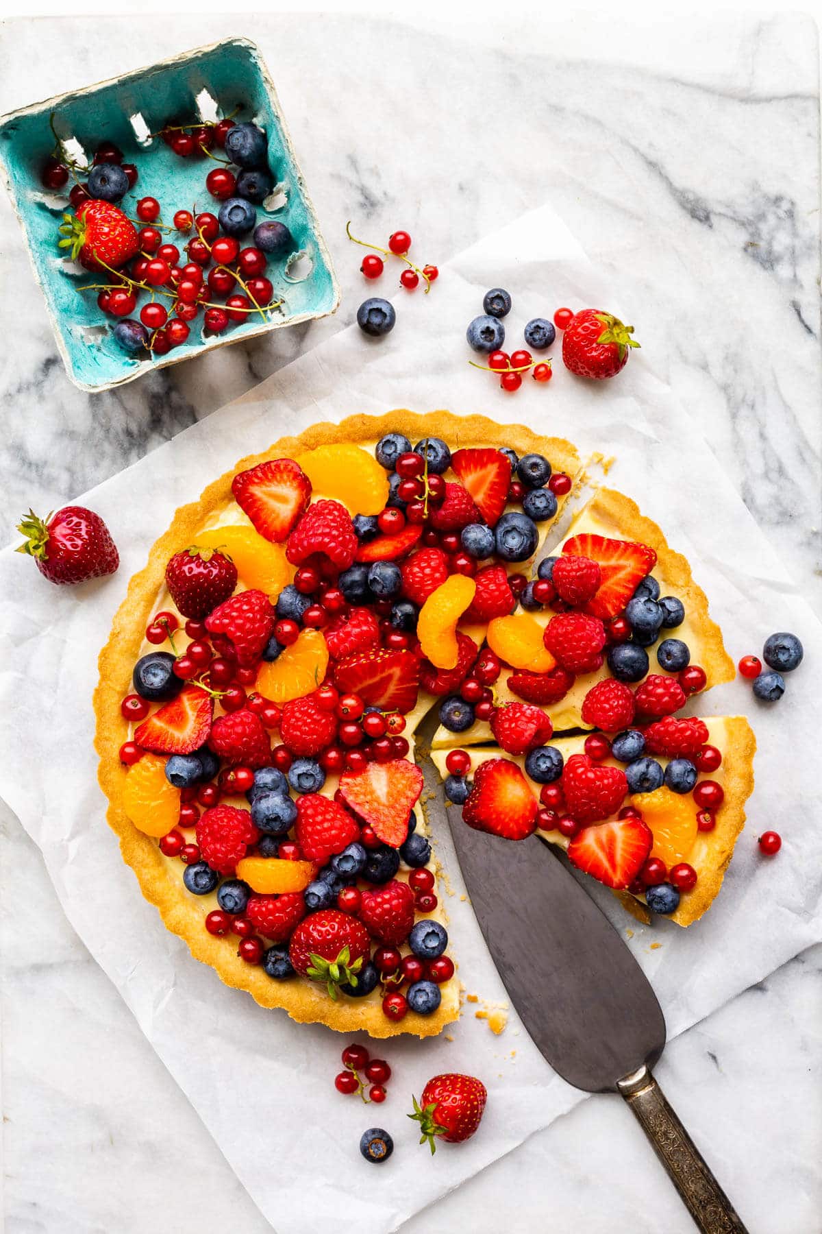 Fruit tart with vanilla pastry cream and fresh berries on parchment paper on marble.