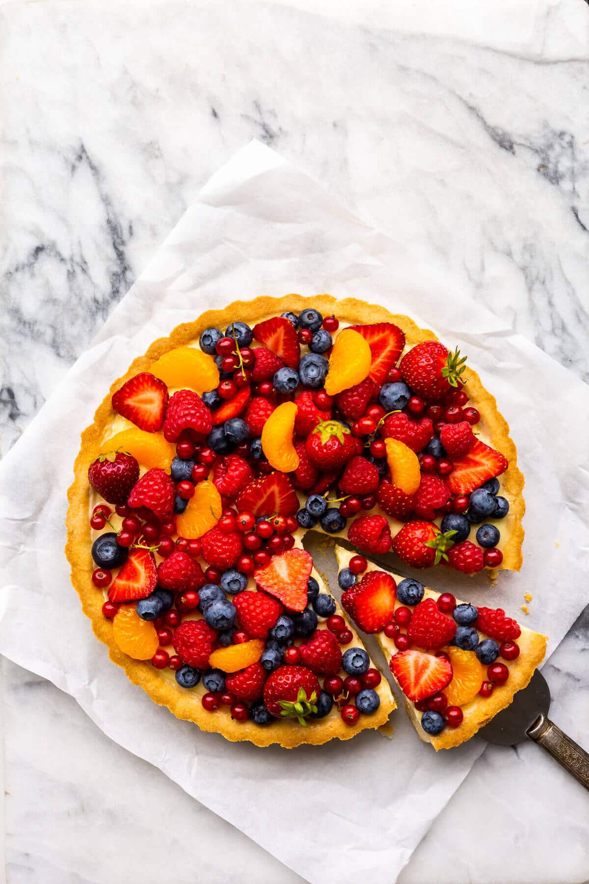 Fruit tart with vanilla pastry cream and fresh berries on parchment paper on marble.