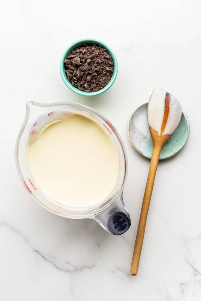 Mint crème anglaise, a wooden spoon, and a bowl of chunks of chocolate, ready to make mint chocolate chip ice cream.