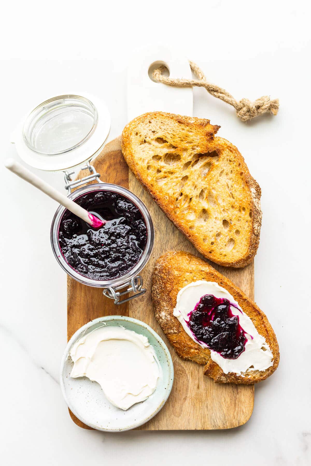 A jar of homemade blueberry jam served with cream cheese and toasted sourdough on a wood board.