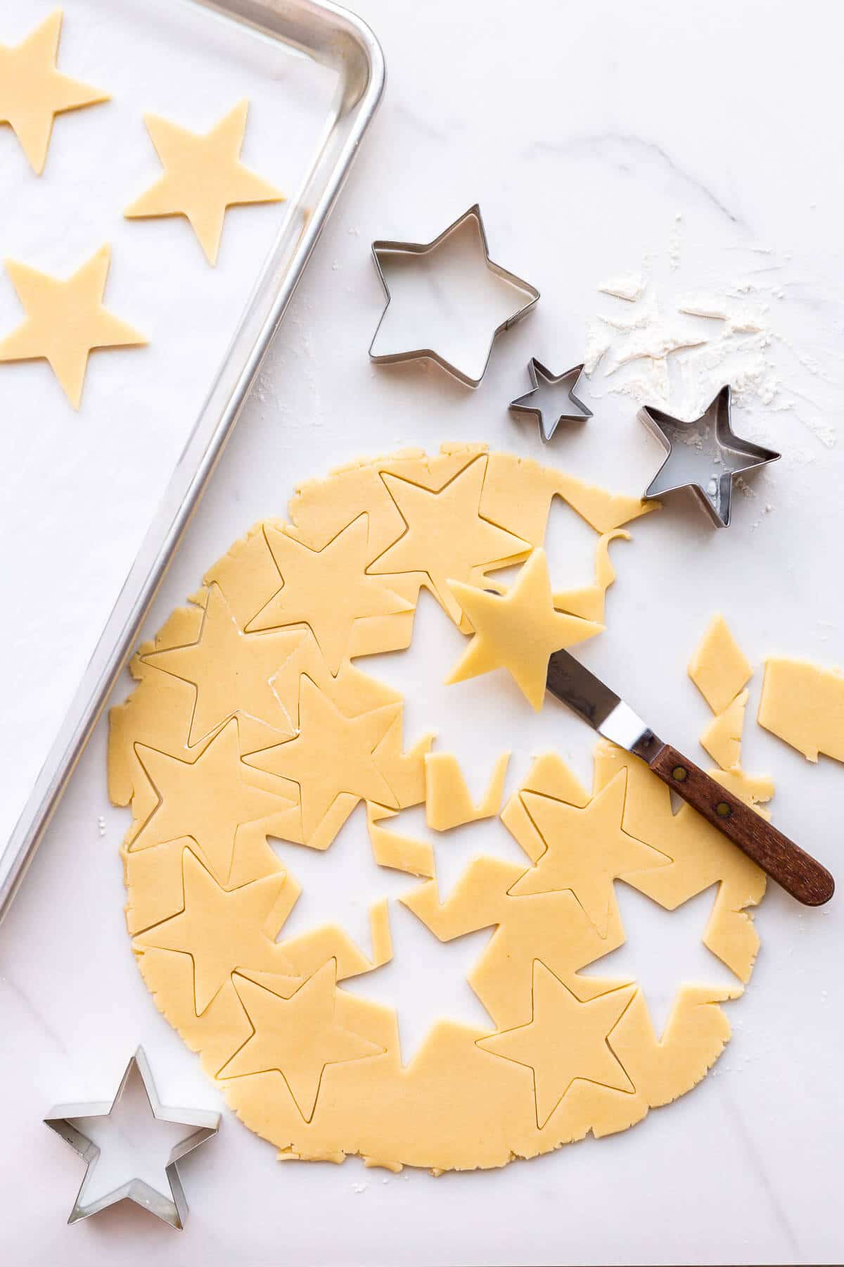 Cutting out stars from rolled sugar cookie dough with cookie cutters before transferring to a sheet pan lined with parchment.