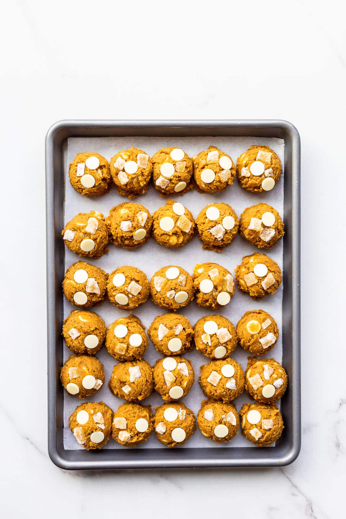 Scoops of ginger cookie dough with white chocolate chips before baking on a parchment-lined sheet pan.