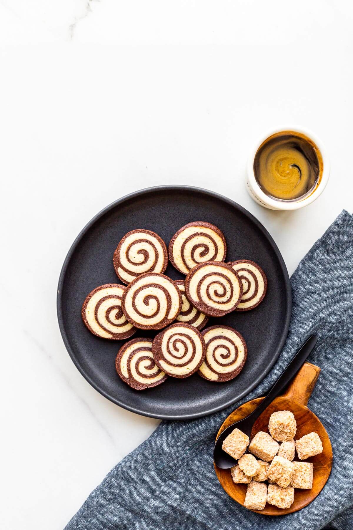 A plate of chocolate pinwheel cookies served with a cup of coffee and a bowl sugar cubes.