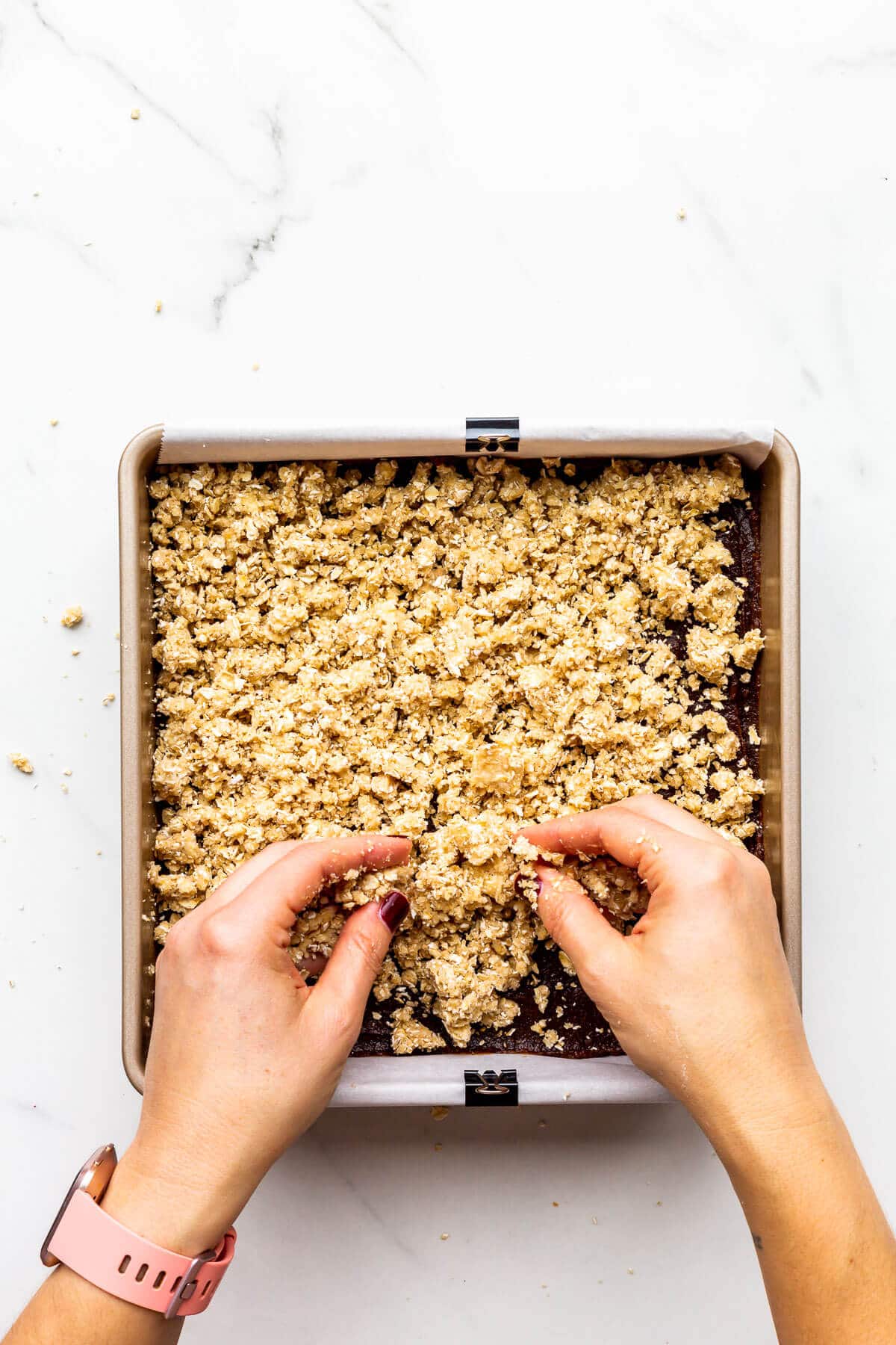 Using hands to crumble oat topping on a pan of date squares before baking in a 9x9 pan.
