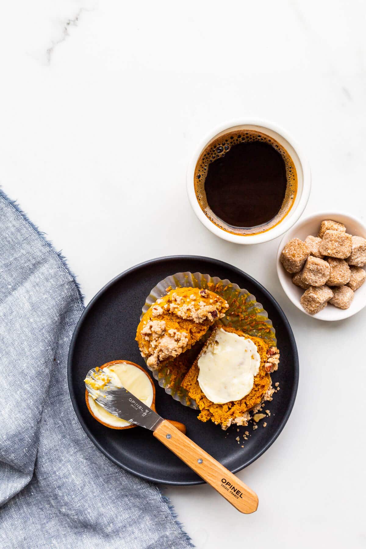 A plate with a pumpkin muffin split open and smeared with butter, with sugar cubes on the side for coffee.