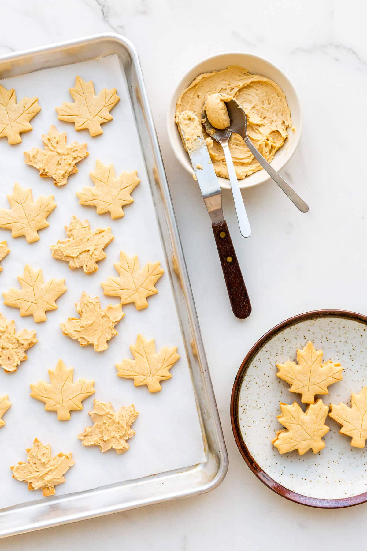 Assembling a sheet pan of maple cookies by spreading a maple cream filling on one maple shortbread cookie, and topping with another.