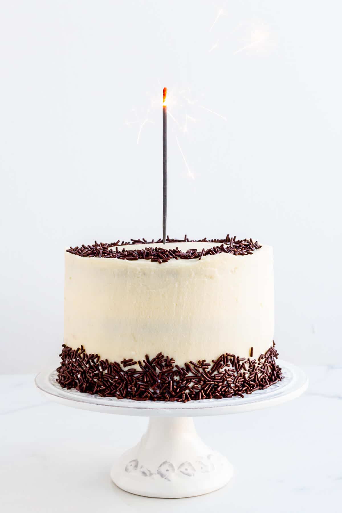 Chocolate layer cake with cream cheese frosting and chocolate sprinkles on a cake stand with a sparkler.