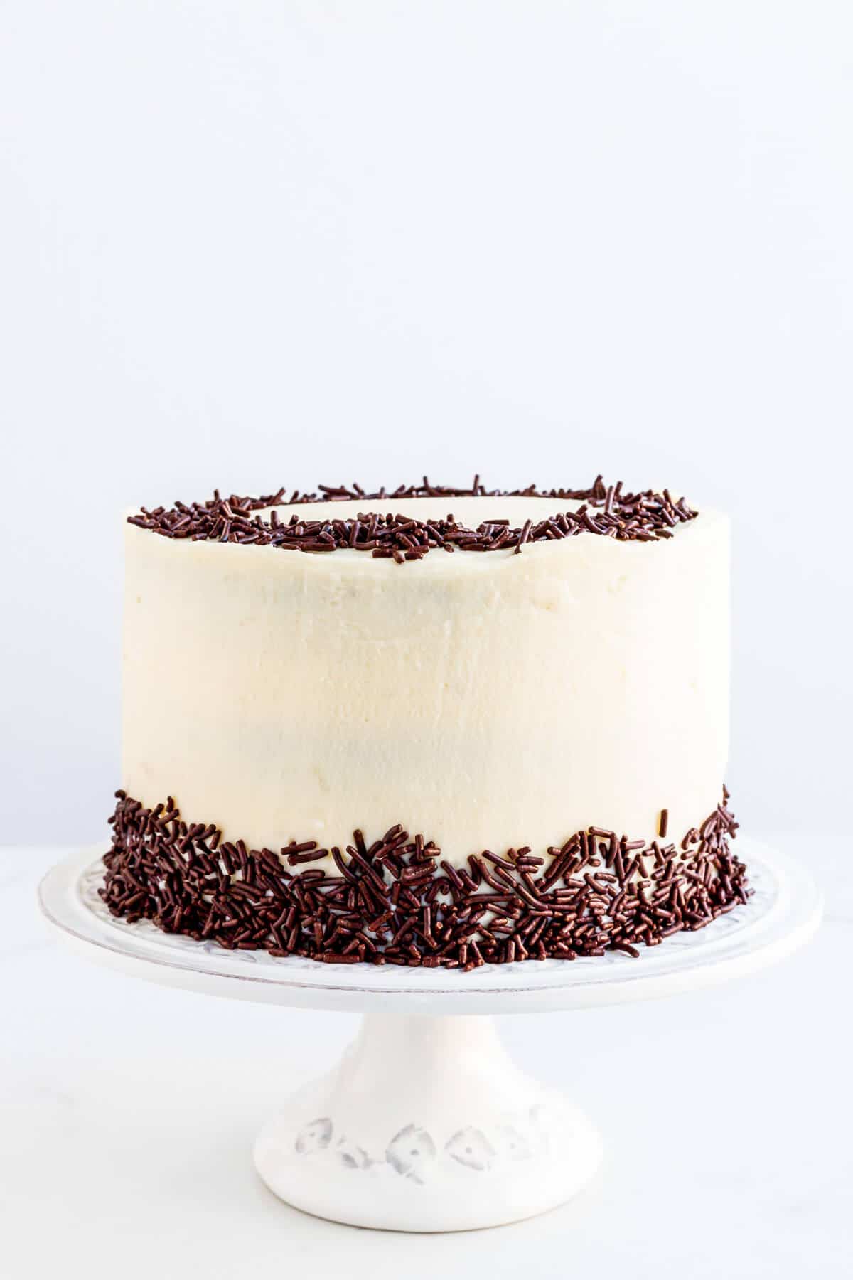 Chocolate layer cake with cream cheese frosting and chocolate sprinkles on a cake stand.