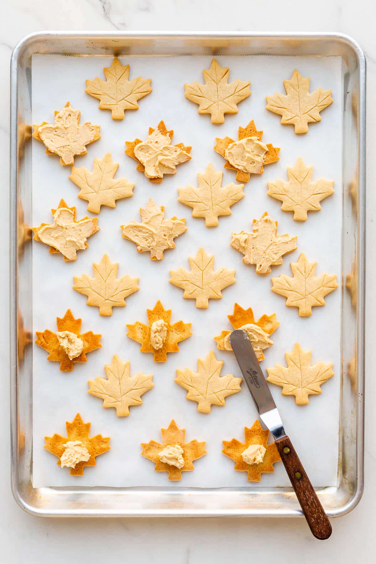 Spreading maple butter filling on maple leaf shortbread cookies with an offset spatula on a sheet pan to make maple cream cookies.