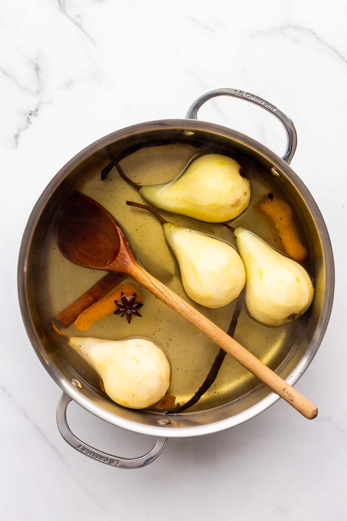 Peeled whole pears in a pot with syrup, spices, and orange peel, ready for poaching on the stove.