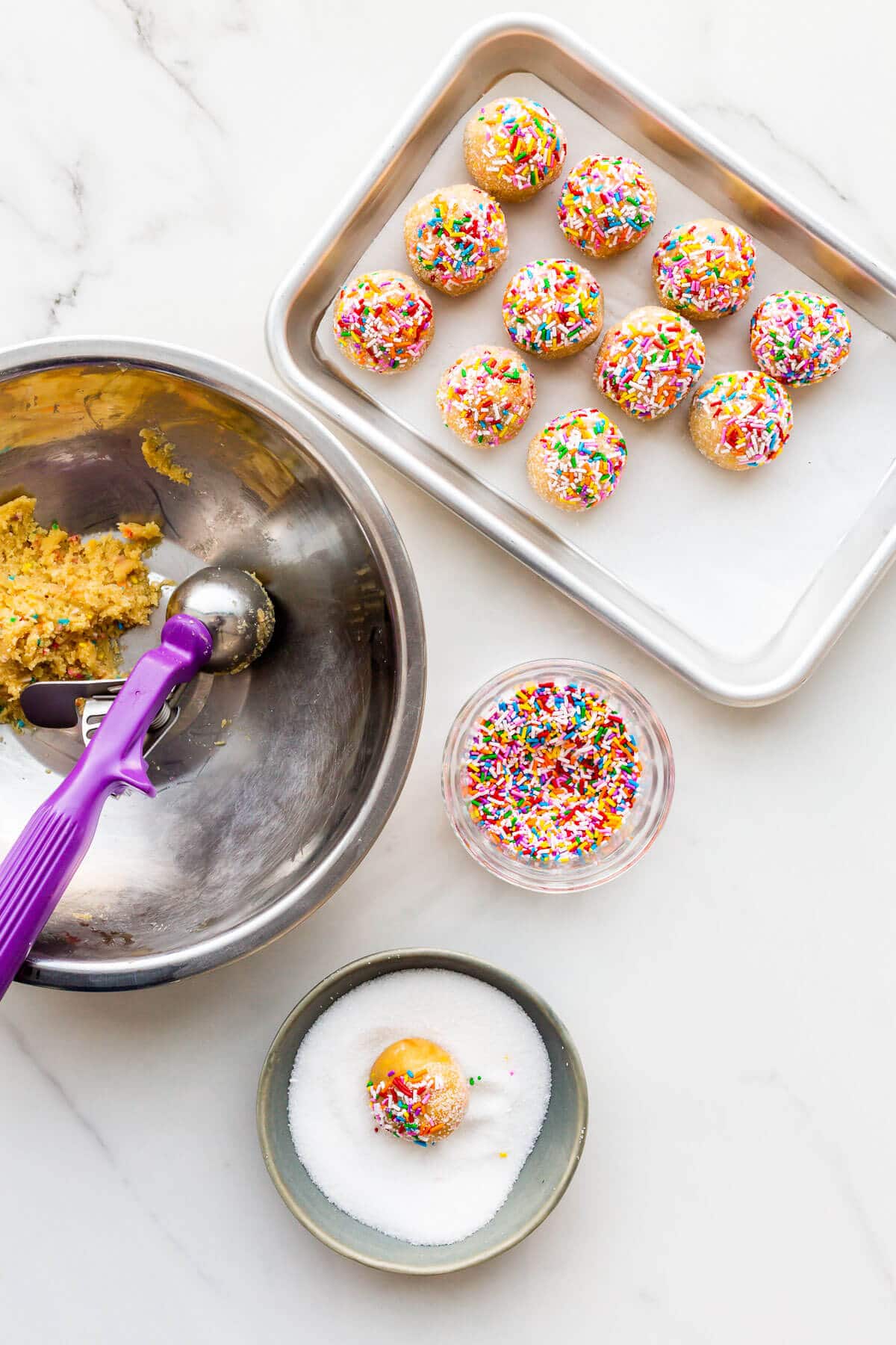 Scooping sugar cookie dough and rolling it in sprinkles and sugar before baking on a sheet pan.