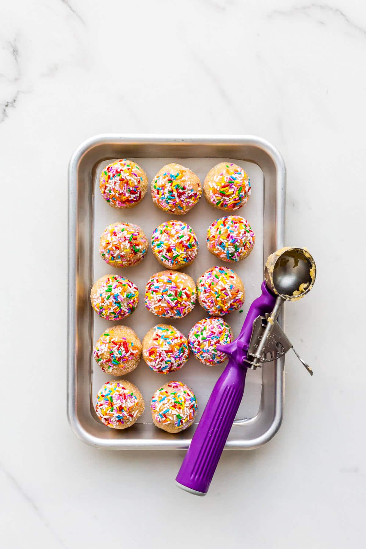 Scooped sugar cookie dough rolled in sprinkles and sugar before baking on a sheet pan.