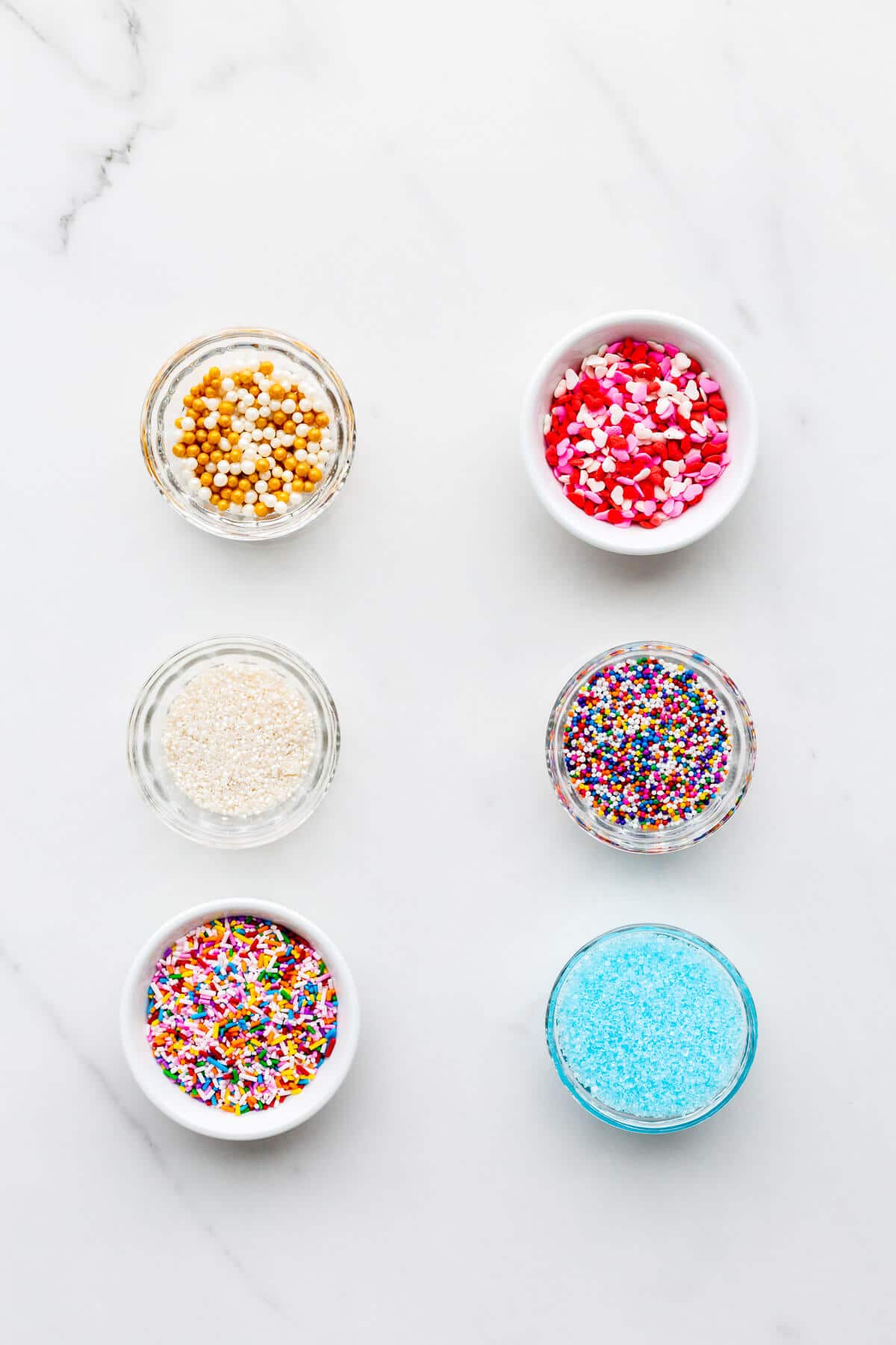 Different types of colourful sprinkles, quins, and sanding sugar in small bowls.