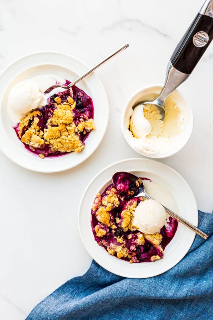 Serving apple and blueberry crumble with scoops of vanilla ice cream on plates.