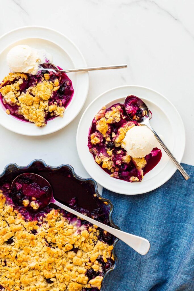 Serving apple and blueberry crumble with scoops of vanilla ice cream on plates.