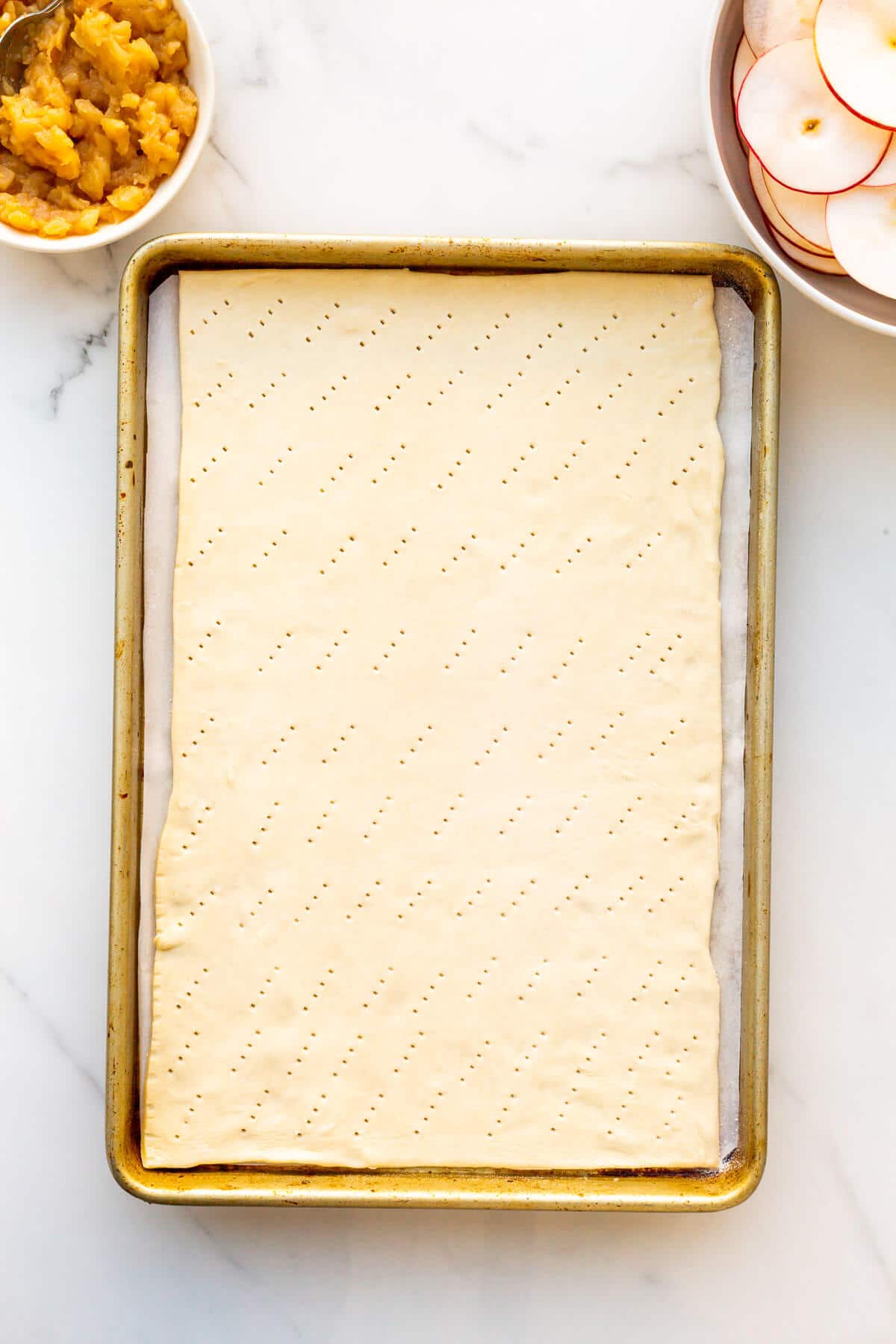 A sheet of puff pastry on a sheet pan, docked with a fork to make air vents to prevent the pastry from bubbling up in the oven.