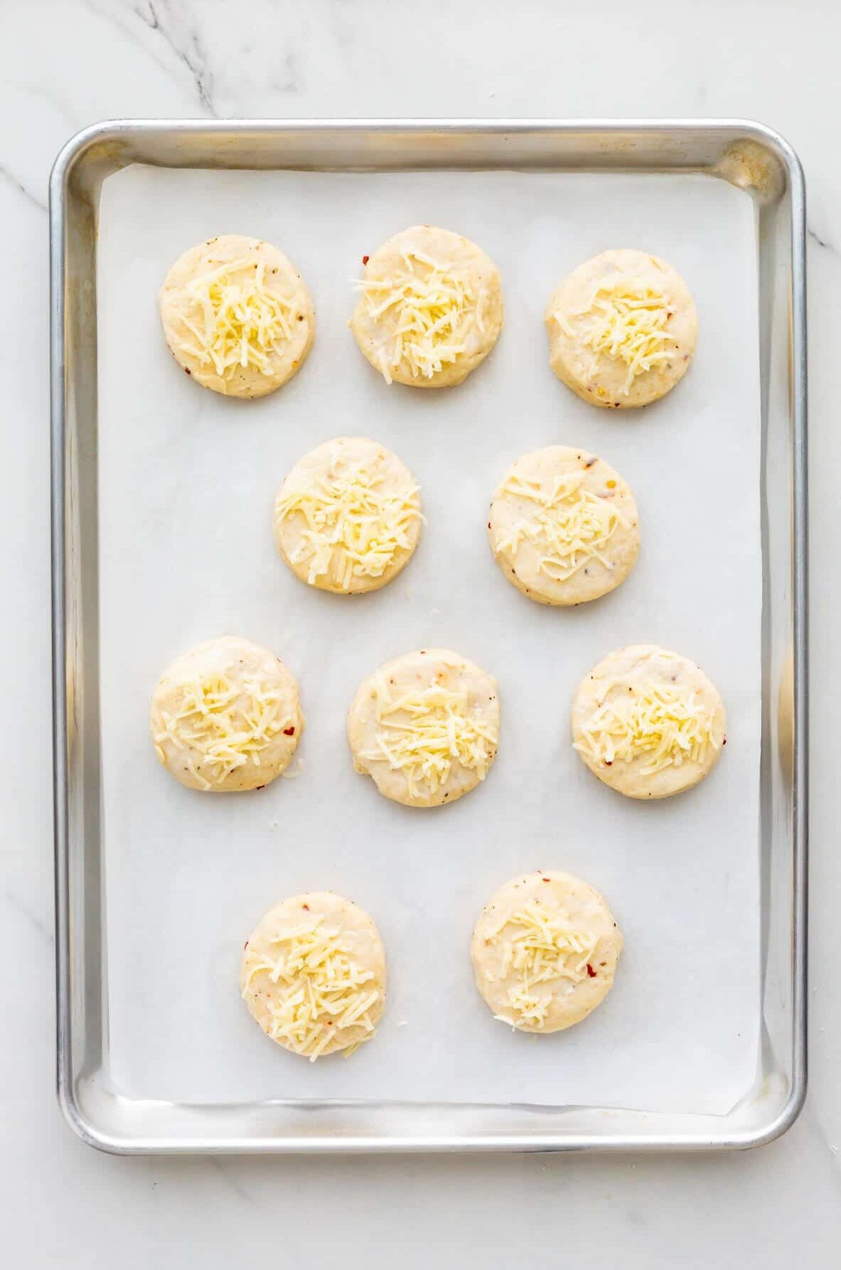 Cheese scones topped with shredded cheese spaced out on a parchment-lined sheet pan before baking.