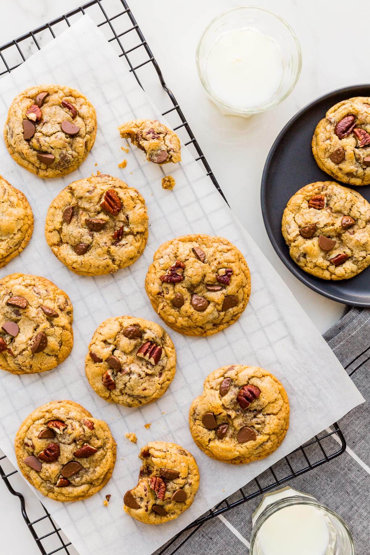 Chocolate chip cookies with pecans and chunks of milk chocolate served from a cooling rack onto a black plate with a glass of milk.