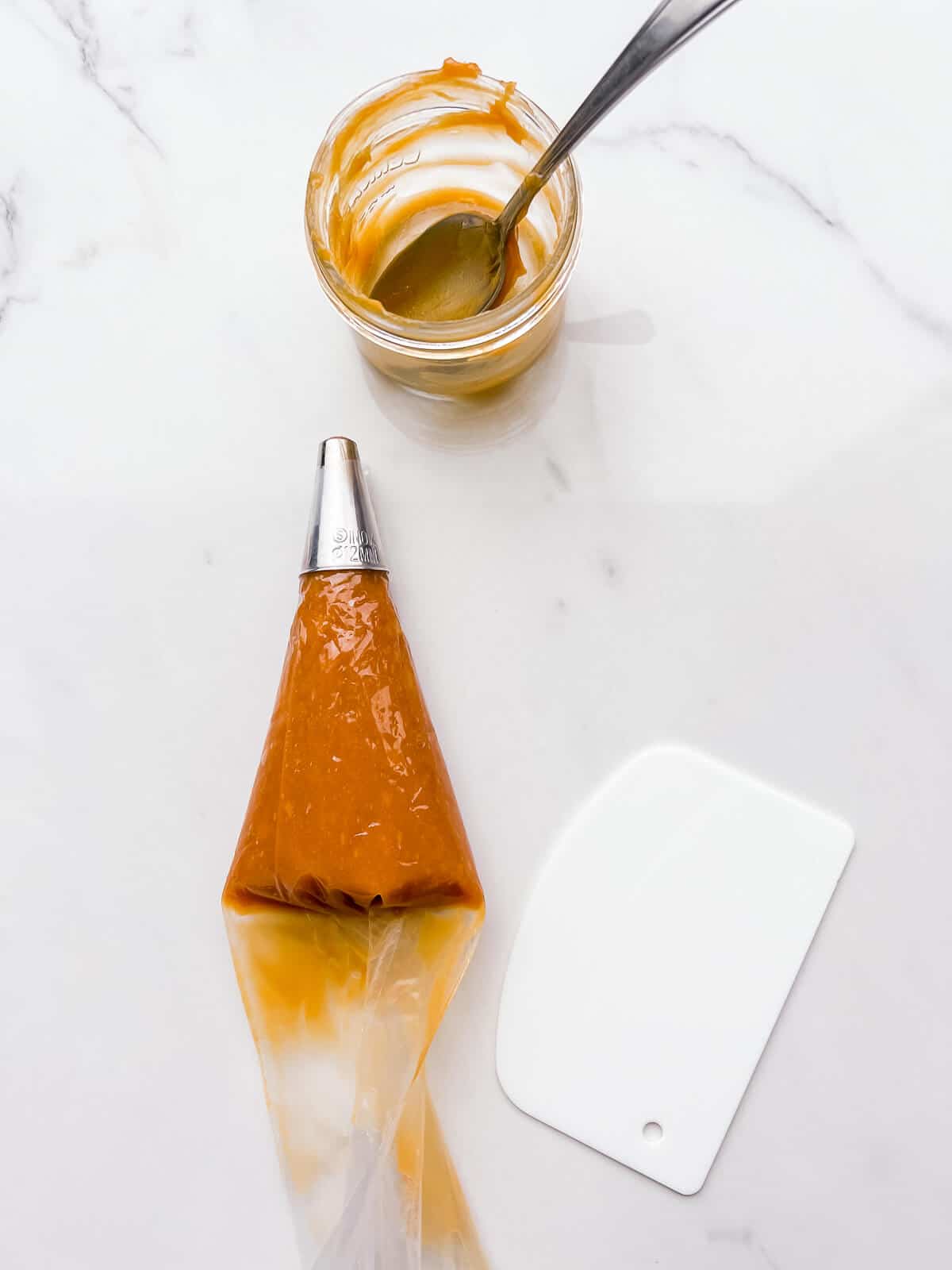 Filling a piping bag with dulce de leche and pushing the filling to the front of the bag with a bench scraper.