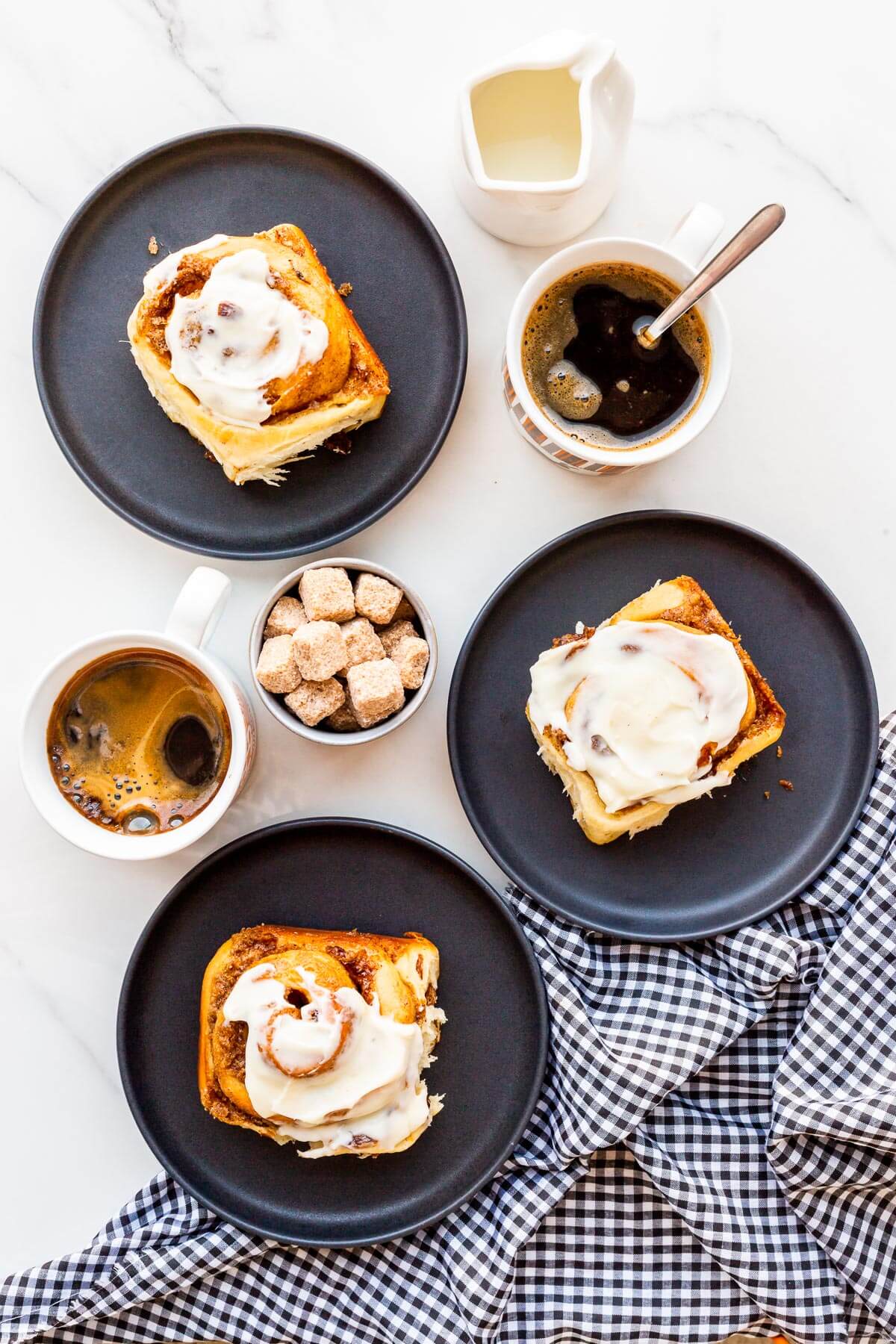 Cinnamon rolls with raisins and cream cheese frosting served on black plates with cups of coffee.