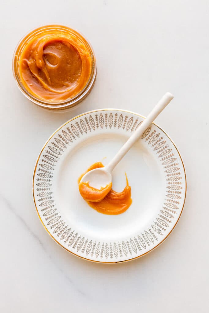 A spoonful of thick, gooey dulce de leche on a plate with the jar it came from on the side.