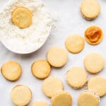 Alfajores cookies filled with dulce de leche being rolled in a bowl of shredded coconut to coat the edges.