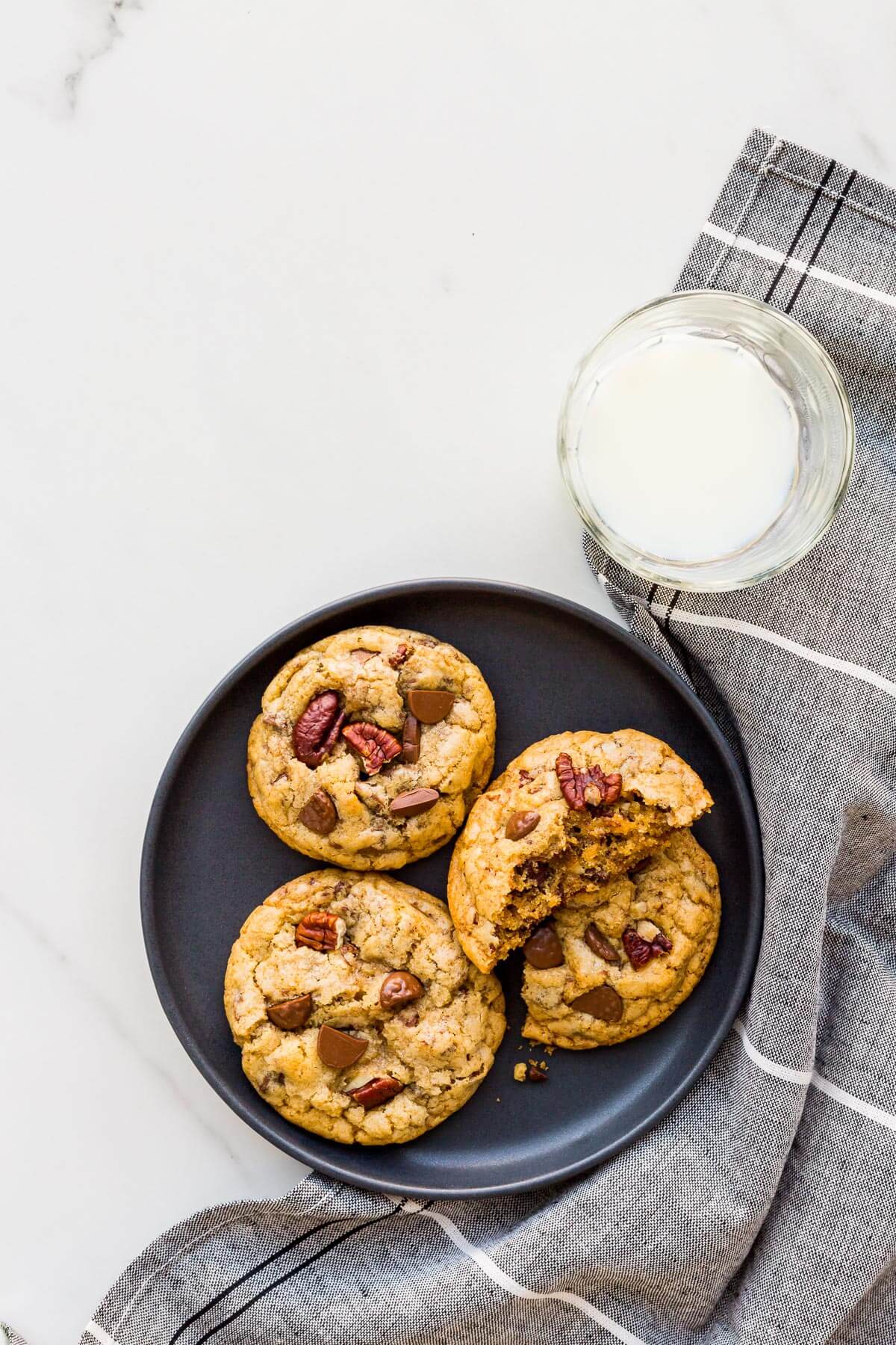 Chocolate chip cookies with pecans and chunks of milk chocolate on a black plate served with a glass of milk.